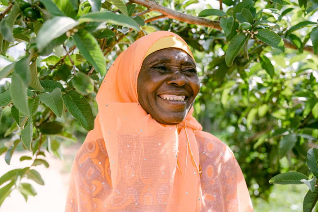 Smiling woman in an orange hijab standing among lush green trees.