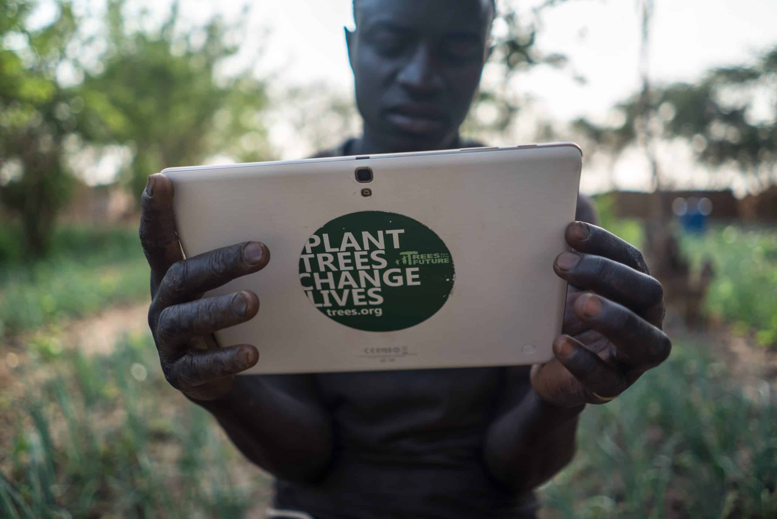 Person holding tablet with 'Plant Trees, Change Lives' sticker, representing Trees for the Future Certification challenge.